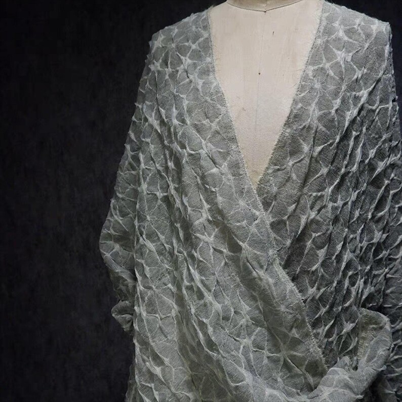 May include: A light gray, textured wrap-style garment with a V-neck design. The fabric has a repeating, geometric pattern, creating a layered effect. The garment is draped over a mannequin, showcasing its shape and texture.