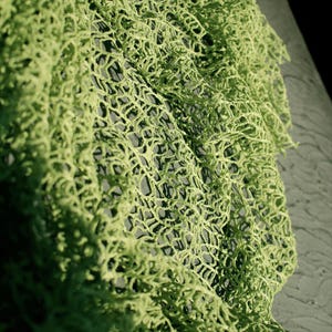 May include: A light green crochet fabric featuring an intricate, open-work mesh pattern with interconnected loops. The textile displays a textured, three-dimensional surface with varying densities of yarn, creating a complex, organic, and web-like structure.