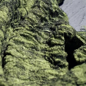 Peut inclure: Gros plan sur un tissu texturé à l'aspect froissé. Le tissu est principalement vert olive avec des accents vert foncé et noirs, créant un aspect organique en couches. La matière semble douce et fluide, avec des crêtes et des plis visibles.