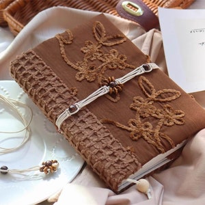 May include: A brown journal with a decorative embroidered cover and white cord closures. The journal is lying on a white surface with a white cord necklace with brown beads.