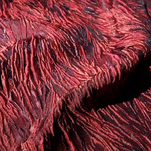 Può includere: Primo piano di un tessuto strutturato di colore rosso intenso con accenti scuri. Il tessuto ha un aspetto increspato e ondulato, con linee verticali e una leggera lucentezza. L'effetto complessivo è ricco e lussuoso, suggerendo un tessuto di alta gamma.