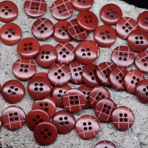 May include: A collection of round, red buttons with four holes. Some buttons have a grid pattern etched on the surface. The buttons are scattered on a textured, gray surface. These buttons are suitable for sewing and craft projects.