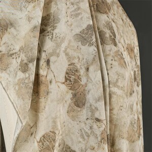 May include: A lightweight fabric scarf featuring an intricate botanical print with overlapping leaf patterns in shades of beige, tan, and muted brown. The material displays soft folds and a semi-sheer texture, highlighting the organic, earth-toned foliage motifs.