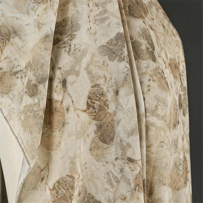 May include: A lightweight fabric scarf featuring an intricate botanical print with overlapping leaf patterns in shades of beige, tan, and muted brown. The material displays soft folds and a semi-sheer texture, highlighting the organic, earth-toned foliage motifs.