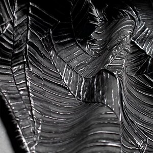 Può includere: Primo piano di un tessuto bianco e nero con un motivo a forma di foglia. Il tessuto ha un aspetto strutturato e plissettato con riflessi bianchi che accentuano i dettagli simili a venature. L'effetto complessivo è un design drammatico ad alto contrasto.