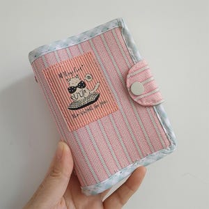Pode incluir: Carteira listrada rosa e branca com um desenho de gato de desenho animado. A carteira tem um fecho de botão e uma borda estampada em azul claro e branco. O gráfico diz "All Things Cute" e "The Wait is for you."