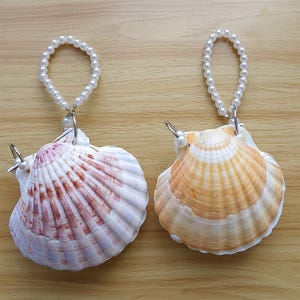 Puede incluir: Dos adornos de conchas marinas con colgadores de perlas. Una concha es blanca y rosa, la otra es blanca y naranja. Ambas conchas tienen un anillo de metal plateado y una pequeña perla.