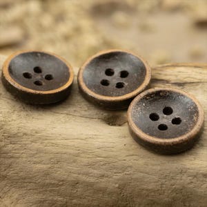 以下が含まれることがあります： 明るい茶色の木材の表面に配置された、それぞれ4つの穴がある3つの濃い茶色のボタン。ボタンはわずかに盛り上がった中央部と、より明るい茶色の縁があります。これらのボタンは、縫製やクラフトプロジェクトに適しています。