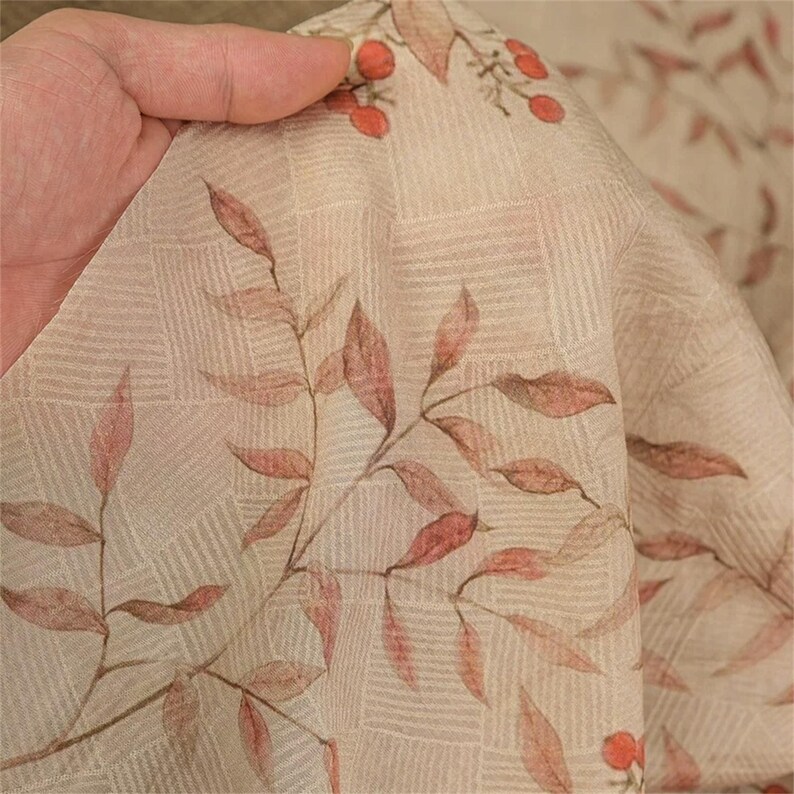 Op de afbeelding: Close-up van een beige stof met een subtiel ruitpatroon. De stof is versierd met een delicate print van roodbruine bladeren en kleine rode bessen, wat een natuurlijk, botanisch thema cre&euml;ert. De stof wordt vastgehouden, wat de textuur en de drapering benadrukt.