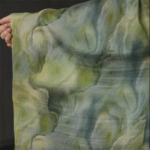 May include: A flowing fabric with a watercolor-like design. The textile features a blend of green, blue, and yellow hues, creating a soft, layered effect. The fabric is held up, showcasing its drape and texture. The overall aesthetic is artistic and ethereal.