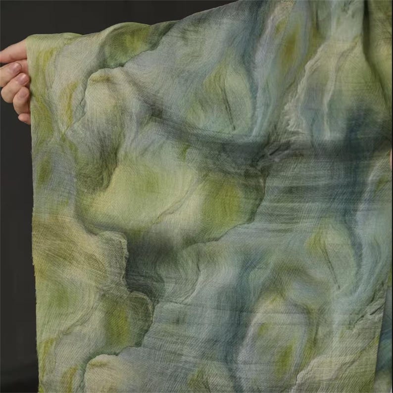 May include: A flowing fabric with a watercolor-like design. The textile features a blend of green, blue, and yellow hues, creating a soft, layered effect. The fabric is held up, showcasing its drape and texture. The overall aesthetic is artistic and ethereal.