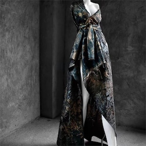 May include: A floor-length, patterned gown in shades of teal, gold, and brown, displayed on a mannequin. The dress features a wrap-style bodice with a tie at the waist and a high slit in the skirt, showcasing a flowing, asymmetrical design.