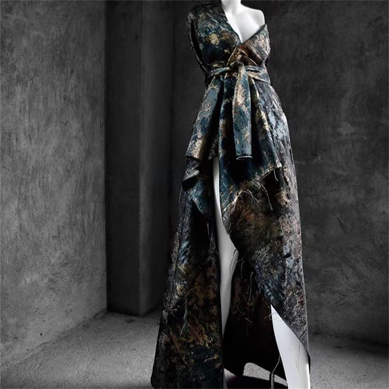 May include: A floor-length, patterned gown in shades of teal, gold, and brown, displayed on a mannequin. The dress features a wrap-style bodice with a tie at the waist and a high slit in the skirt, showcasing a flowing, asymmetrical design.