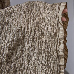 May include: Close-up of a textured fabric in shades of beige and brown. The fabric has a crinkled, irregular surface, with a ruffled edge. The material appears to be a woven textile, possibly linen or a similar natural fiber. The overall aesthetic is earthy and organic.