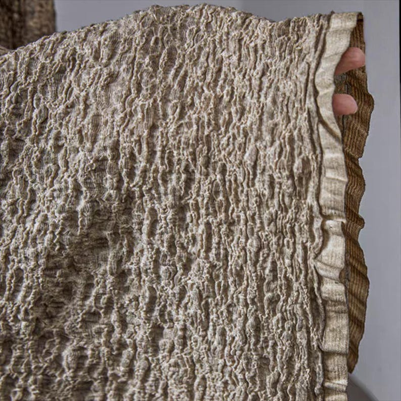 May include: Close-up of a textured fabric in shades of beige and brown. The fabric has a crinkled, irregular surface, with a ruffled edge. The material appears to be a woven textile, possibly linen or a similar natural fiber. The overall aesthetic is earthy and organic.