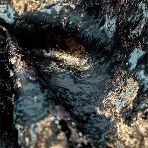 May include: Close-up of a textured fabric with a dark teal, black, and gold color scheme. The fabric has a swirling pattern with metallic gold accents, creating a luxurious and elegant appearance. The fabric appears to be draped.