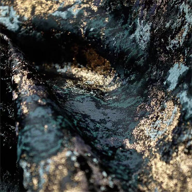 May include: Close-up of a textured fabric with a dark teal, black, and gold color scheme. The fabric has a swirling pattern with metallic gold accents, creating a luxurious and elegant appearance. The fabric appears to be draped.
