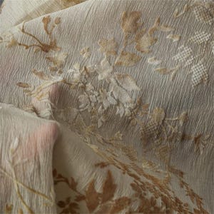 Puede incluir: Primer plano de una tela beige con una superficie texturizada y un estampado floral en tonos marrones y beige. La tela está doblada, revelando la textura y los detalles del diseño. El estampado floral incluye hojas y flores.