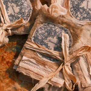 May include: Close-up of several handmade journals. Each journal features a burlap cover with a floral design and the word "Journal" printed on a circular label. The journals are tied with cream-colored ribbons and have decorative stitching.