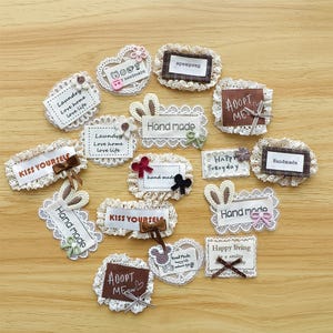 Könnte beinhalten: Eine Sammlung von Stoffaufnähern mit verschiedenen Texten und dekorativen Elementen. Die Aufnäher zeigen Sätze wie "Hand made", "Kiss Yourself" und "Adopt Me". Sie sind mit Spitze beschnitten und mit Schleifen und kleinen Verzierungen versehen.
