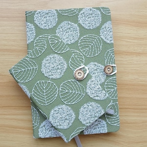 Peut inclure: Un journal en tissu vert avec des motifs de feuilles et de cercles brodés en blanc. Le journal a une couverture assortie avec une fermeture à bouton. Un signet en ruban gris est visible.