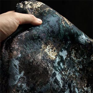 May include: Close-up of a dark fabric with a textured, marbled appearance. The fabric features a blend of black, teal, and gold metallic accents, creating a luxurious and artistic look. The material is likely used for apparel or home decor.