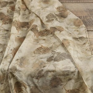 May include: A lightweight fabric scarf featuring an all-over botanical print with various leaf shapes in shades of muted brown, tan, and soft beige. The textile displays a natural, organic pattern with overlapping foliage silhouettes across the surface.