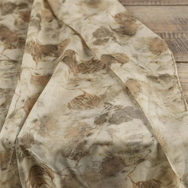 May include: A lightweight fabric scarf featuring an all-over botanical print with various leaf shapes in shades of muted brown, tan, and soft beige. The textile displays a natural, organic pattern with overlapping foliage silhouettes across the surface.