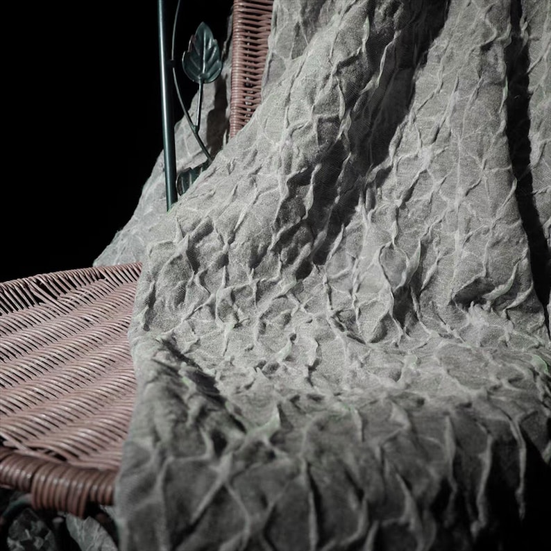 May include: A gray, textured fabric draped over a wicker chair. The fabric has a repeating diamond pattern and a soft, wrinkled appearance. The chair has a metal back with a leaf design.