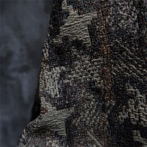 May include: Detailed view of a textured fabric with a woven design. The material displays a blend of dark brown, grey, and olive green hues, resulting in a mottled effect. The weave is clearly visible, providing depth and texture to the fabric.