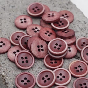 May include: A collection of round, burgundy buttons with four holes each. The buttons have a slightly lighter, off-white rim, and are scattered on a textured, gray surface. These buttons are ideal for sewing and craft projects.