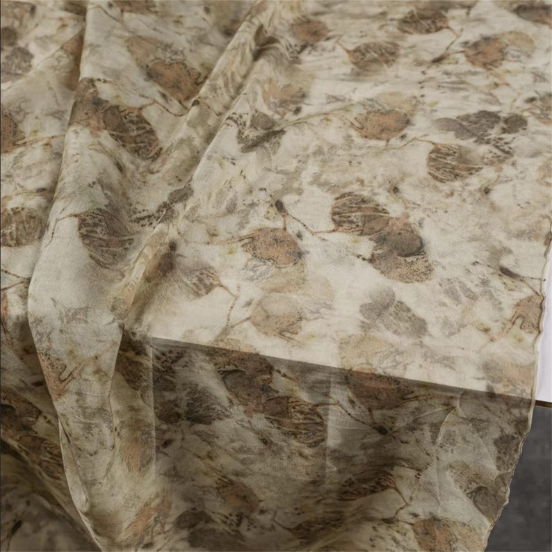May include: A lightweight fabric featuring an all-over botanical print with overlapping leaf silhouettes in shades of muted brown, tan, and cream. The textile displays a semi-sheer quality with a soft, draped texture and visible organic patterns.