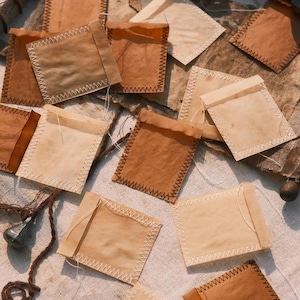 Op de afbeelding: Een verzameling vierkante papieren zakjes in diverse tinten bruin en beige. Elk zakje is voorzien van zichtbare witte zigzagstiksels langs de randen en heeft een dun wit draadje aan de bovenkant, wat zorgt voor een textuurrijk uiterlijk.