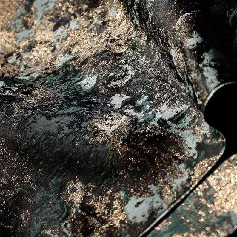 May include: Close-up of a textured fabric with a dark teal, black, and gold metallic design. The fabric has a marbled appearance with areas of light reflection, creating a luxurious and abstract pattern. The material appears to be soft and flowing.
