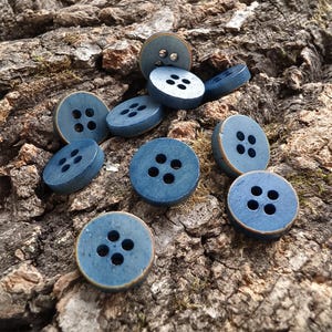 May include: A collection of round, blue buttons, each featuring four holes arranged in a square pattern. The buttons have a smooth, matte finish with subtle, worn-looking edges that reveal a lighter, brownish tone along the outer rim.