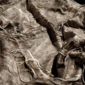 Può includere: Primo piano di un tessuto marrone strutturato con un aspetto invecchiato. Il tessuto ha una trama intrecciata con fili visibili e un bordo leggermente sfilacciato. Il colore è un marrone tenue con luci e ombre.