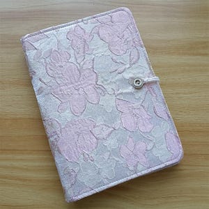 May include: A floral-patterned notebook cover in shades of pink and white. The cover features a repeating design of large flowers and a button closure. The notebook is closed and appears to be made of a textured fabric.