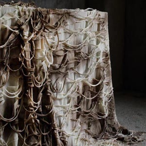 Può includere: Un tessuto drappeggiato nei toni del crema e del marrone, con un design strutturato e stratificato. Il tessuto presenta elementi ad anello e arricciati, creando un effetto tridimensionale. L'estetica generale è morbida e organica.