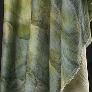 May include: Close-up of draped fabric in shades of green and cream. The fabric has a watercolor-like pattern with swirling, organic shapes. The texture appears soft and lightweight, with a slightly frayed edge visible.