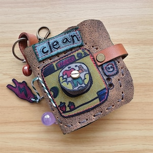 May include: Handmade brown leather wallet with a colorful, artistic design. The wallet features a small, green patch with the word "clean" embroidered in blue. Additional details include a key ring, a leather strap, and decorative stitching.