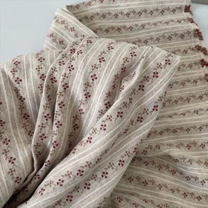 Puede incluir: Tela beige con rayas blancas verticales y pequeños patrones florales en burdeos y marrón. La tela está doblada, mostrando la textura y el diseño. Ideal para proyectos de costura o decoración del hogar.