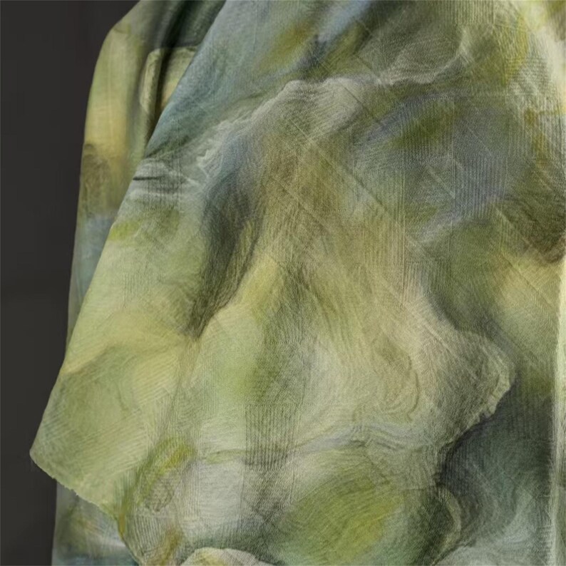 May include: Close-up of a flowing fabric with a watercolor-like design. The fabric features a blend of green, yellow, and blue hues, creating a soft, textured appearance. The material appears lightweight and draped, suggesting a garment or textile.