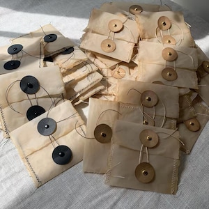 Peut inclure: Un ensemble de petites enveloppes carrées en papier de différentes tailles. Chaque enveloppe est de couleur beige clair et fermée par un bouton et une ficelle. Certaines enveloppes ont des boutons noirs, d'autres des boutons dorés. Les bords sont grossièrement cousus.