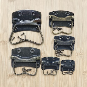 Puede incluir: Un surtido de manijas de maleta de metal de color bronce antiguo con tornillos a juego. Las manijas varían en tamaño y presentan un diseño con bisagras y un asa curva. El hardware se muestra sobre una superficie de madera de color marrón claro.