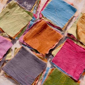 May include: Close-up of a patchwork of colorful fabric squares. The squares are in shades of blue, pink, orange, green, and purple. The edges of the fabric are frayed, giving a handmade, rustic appearance. The fabric appears to be cotton or linen.