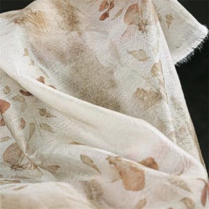 May include: Close-up of a sheer fabric with a delicate leaf pattern. The fabric is primarily off-white with brown and tan leaf designs. The edges of the fabric are frayed, adding a textured element to the design.