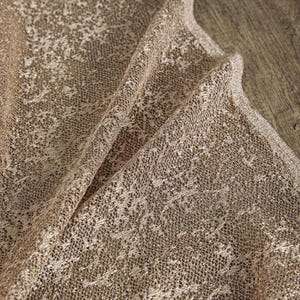 May include: Close-up of a folded, sheer fabric with a delicate, open weave. The fabric is a warm, metallic gold color, with a subtle sheen. The edges are finished with a fine, metallic trim. The overall texture is soft and flowing.