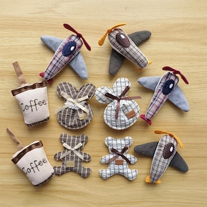 May include: A collection of handmade fabric toys, including airplanes, teddy bears, and bunny-shaped ornaments. The airplanes have plaid wings and propellers. The coffee cups have the word "Coffee" printed on them. The toys are arranged on a wooden surface.