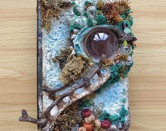 Könnte beinhalten: Ein handgefertigtes Mixed-Media-Kunstwerk mit einem großen, runden Glaselement. Das Kunstwerk ist mit natürlichen Elementen wie Moos, kleinen Kürbissen und Ästen verziert, wodurch eine skurrile, von der Natur inspirierte Ästhetik entsteht. Die Farbpalette umfasst Braun-, Grün- und Blautöne.