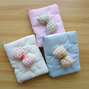 May include: Three quilted pot holders in pastel colors: pink, blue, and cream. Each has a star pattern and a bow accent in a coordinating gingham pattern. The pot holders have a loop for hanging.